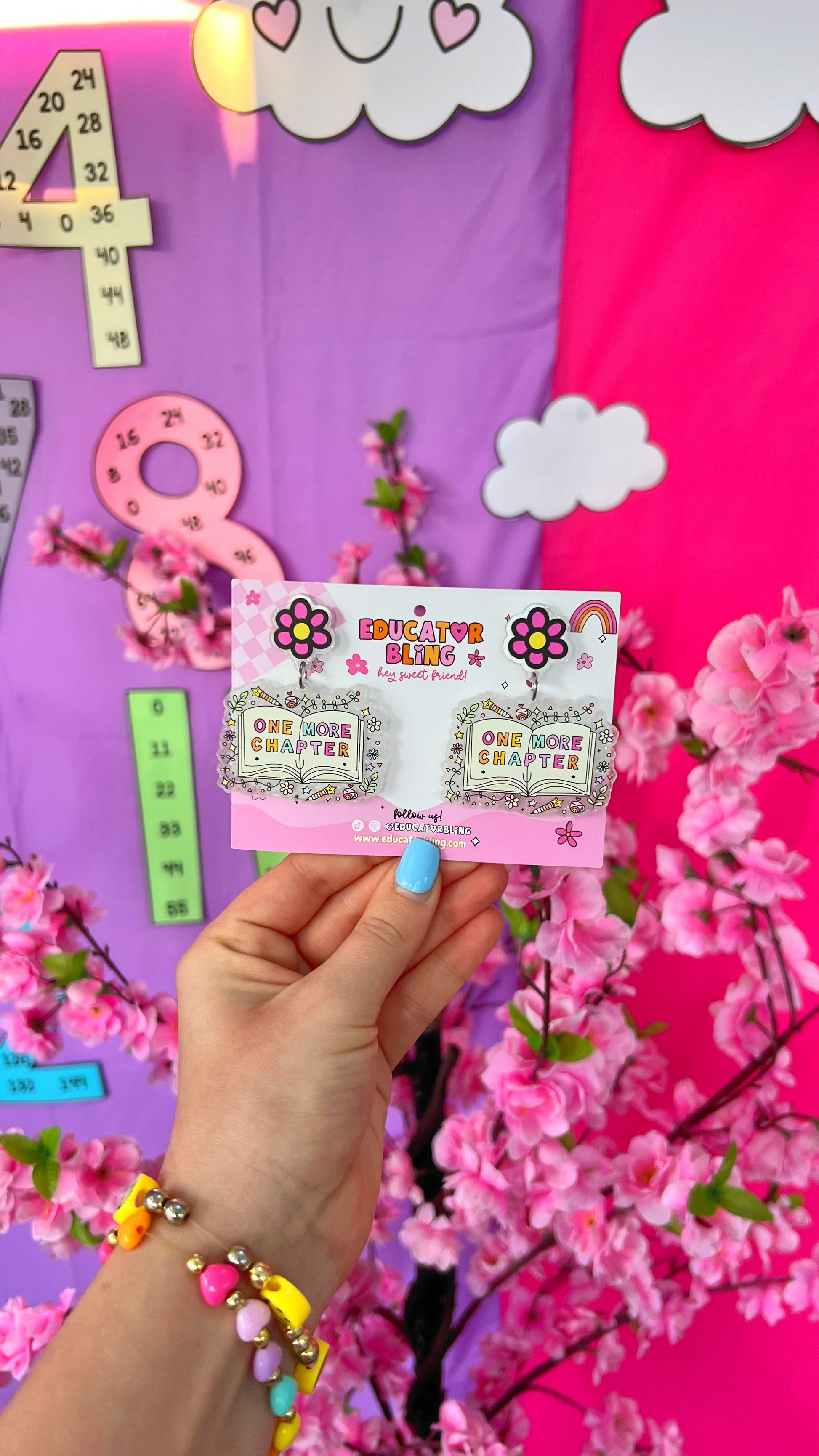 One More Chapter teacher earrings by Educator Bling – colourful acrylic dangle earrings with a pink flower stud and an open book reading 'One More Chapter', on a rainbow display card