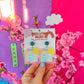 Total Book Worm teacher earrings by Educator Bling – colourful acrylic dangle earrings of a smiling sun and glasses-wearing bookworm hugging a stack of books, on a rainbow display card