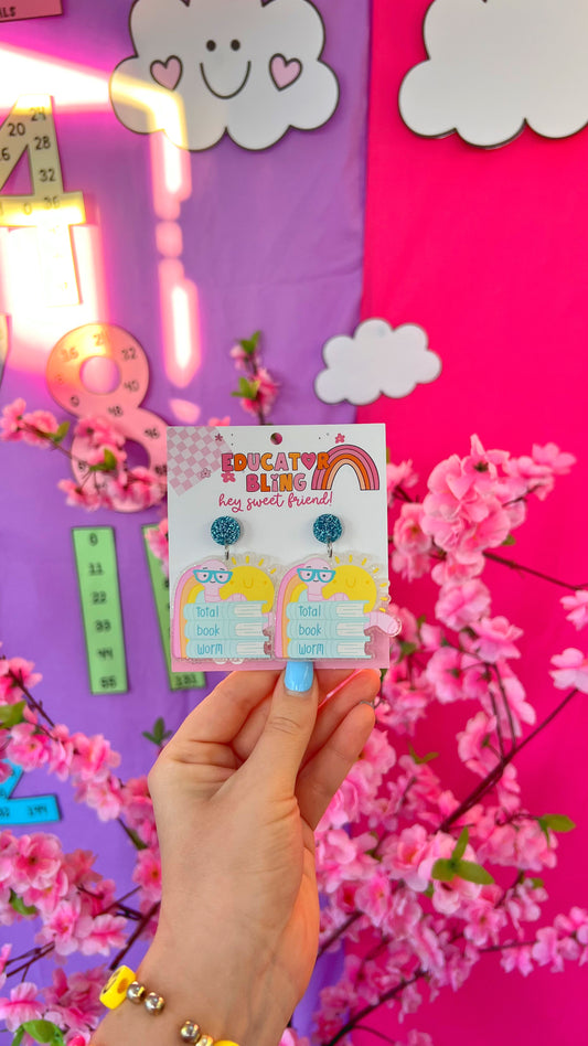 Total Book Worm teacher earrings by Educator Bling – colourful acrylic dangle earrings of a smiling sun and glasses-wearing bookworm hugging a stack of books, on a rainbow display card